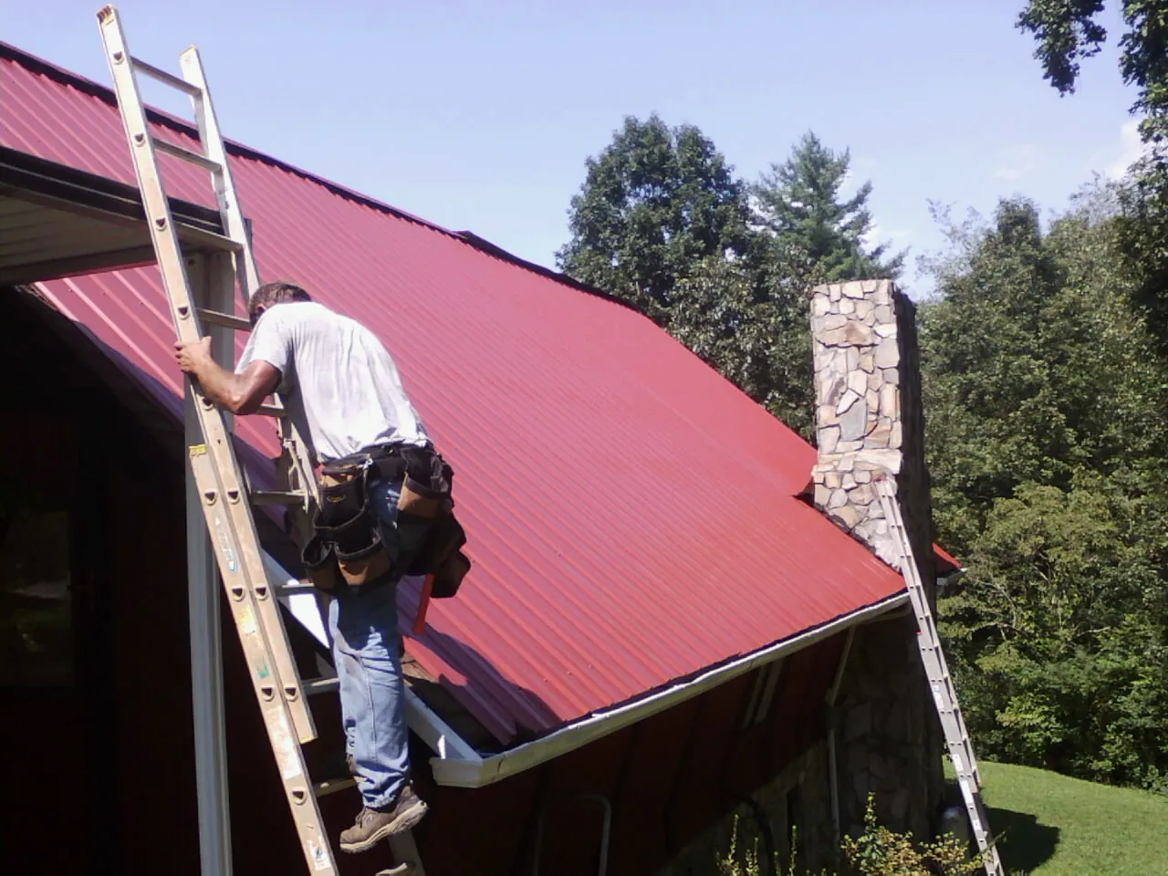Roofer on a ladder beside a red metal roof and stone chimney — Soffit & Fascia Repair by Roofing Brothers in Pottsboro